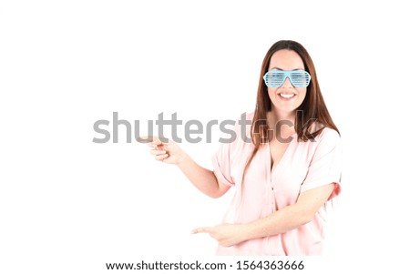Confident young woman smiling and pointing her fingers to one side while wearing blue sunglasses against a white background