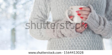 Young woman with morning cup of coffee looking through the window, blurry winter snowy woods outside.