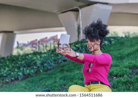 Beautiful brunette with afro hair takes a selfie photo with her tablet sitting in a park