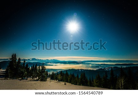 Mesmerizing landscape of snowy ski slope on the backdrop of spruce forest and mountain ranges in the moonlight and blue sky on a clear frosty winter evening. Concept of outdoor recreation in winter