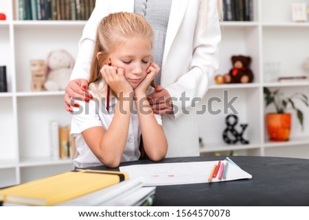 Sad girl sits at the table, draws, her mom hugs her shoulders and comforts