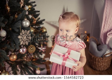 Beautiful little girl with blue eyes in a pink dress holds a gift near the Christmas tree. Christmas. New Year. Holidays.