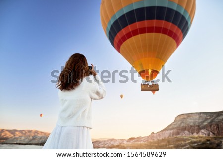 A tourist photographer girl wearing white sweater on a mountain top enjoying wonderful view and taking a picture of balloons in Cappadocia