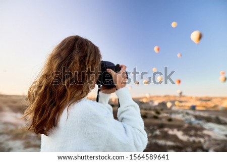A tourist photographer girl wearing white sweater on a mountain top enjoying wonderful view and taking a picture of balloons in Cappadocia