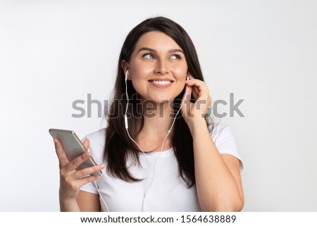 Positive Woman In Earphones Listening To Music On Smartphone Standing On White Background. Studio Shot