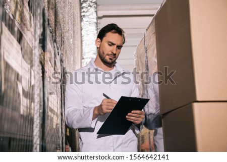 concentrated storekeeper in white coat writing on clipboard in warehouse Royalty-Free Stock Photo #1564642141