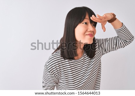 Young beautiful chinese woman wearing black striped t-shirt over isolated white background very happy and smiling looking far away with hand over head. Searching concept.