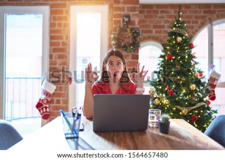 Beautiful woman sitting at the table working with laptop at home around christmas tree looking surprised and shocked doing ok approval symbol with fingers. Crazy expression