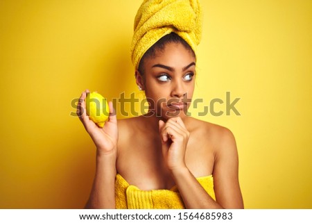 Afro american woman wearing towel after shower holding lemon over isolated yellow background serious face thinking about question, very confused idea