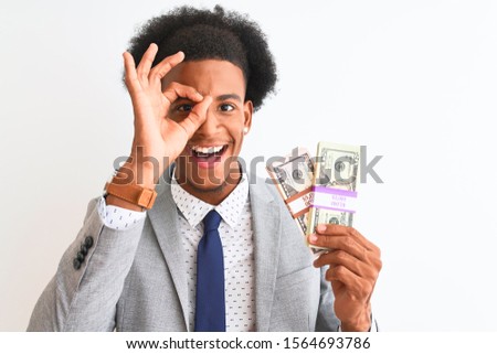 Young african american businessman holding dollars standing over isolated white background with happy face smiling doing ok sign with hand on eye looking through fingers