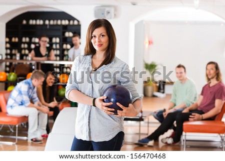 Portrait of confident young woman holding bowling ball with friends in background