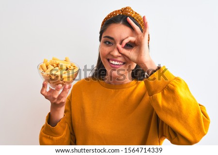Young beautiful woman holding bowl with macaroni pasta over isolated white background with happy face smiling doing ok sign with hand on eye looking through fingers