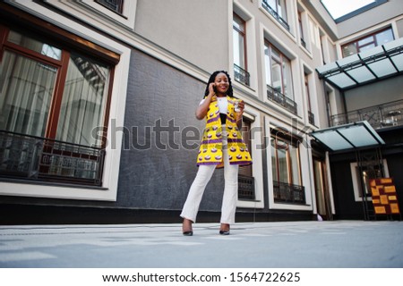 Stylish african american women in yellow jacket posed on street with hot drink in disposable paper cup and mobile phone at hands.