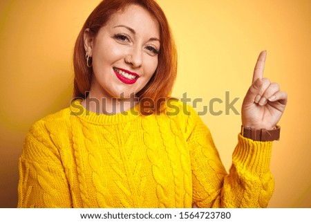 Young redhead woman wearing winter sweater standing over isolated yellow background with a big smile on face, pointing with hand and finger to the side looking at the camera.