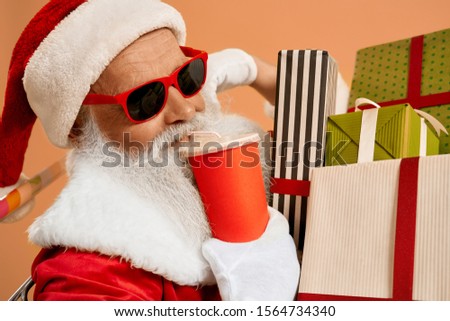 Happy senior man in red traditional Santa costume and stylish sunglasses, drinking soda from paper cup with colorful present boxes around. Santa Claus preparing for giving gifts for happy children.