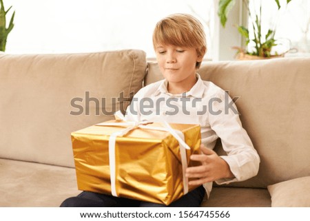 Cute Caucasian teenager sitting on sofa with New Year's gift on his lap. Handsome boy ready to open golden box with Christmas present in it, having curious anticipated facial expression, smiling