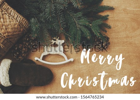 Merry Christmas text sign on reindeer toy, gloves, basket with fir branches and cones on rustic wooden background. Flat lay. Zero waste. Season's greeting card