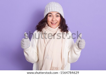 Winter girl with dark wavy hair dresses white warm sweater, pale pink scarf, cap and gloves gesturing thumbs up isolated over lilac background, looks excited, keeping mouth opened, looking at camera,