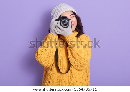 Winter girl looking through photo camera over lilac background, female viewfinder wearing warm knitted yellow sweater, gloves and cap, spending her free time to enjoy her hobby, making photos.
