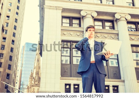 Power of Technology. Young businessman traveling, working in New York City, wearing blue suit, white shirt, standing on street outside office building, working on laptop computer, talking on phone.