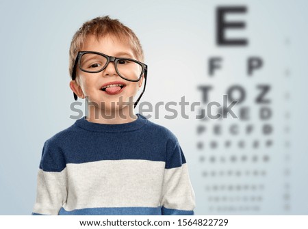 vision and children concept - portrait of smiling little boy with crookedly worn glasses showing tongue over eye test chart background Royalty-Free Stock Photo #1564822729