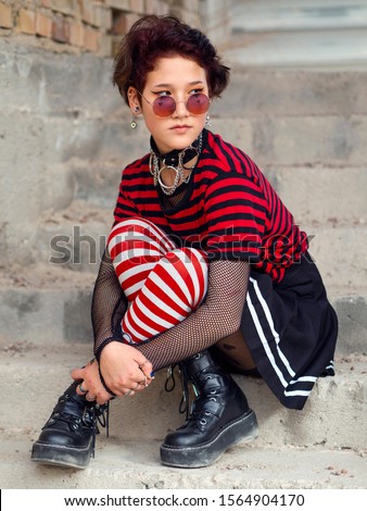 Redhair urban punk girl in a pink hipster sunglasses on a graffiti wall background with copy space. Rebel Goth young woman sitting on the steps