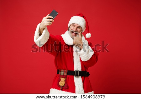 Elderly gray-haired mustache bearded Santa man in Christmas hat isolated on red background. New Year 2020 celebration concept. Mock up copy space. Doing selfie shot on mobile phone, showing thumb up