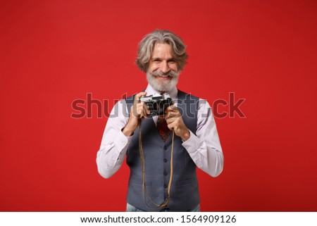 Smiling elderly gray-haired mustache bearded man in classic shirt vest colorful tie isolated on red background in studio. People lifestyle concept. Mock up copy space. Hold retro vintage photo camera