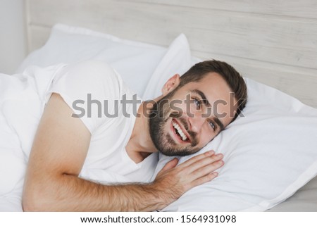 Calm young handsome bearded man lying in bed with white sheet pillow blanket in bedroom at home. Smiling beauty male spending time in room. Rest relax good mood lifestyle concept. Mock up copy space