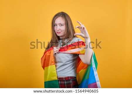 Portrait of concerned young woman in vivid casual clothes looking camera, gesturing demonstrating size with workspace, covered LGBT flag. People lifestyle concept.