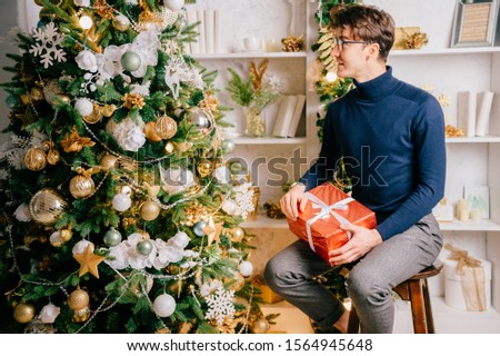 Handsome elegant man holding gift box and and looking at christmas tree in cozy room with new year decorations