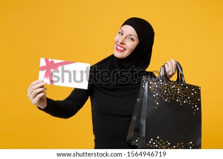 Arabian muslim woman in black hijab hold package bag, purchases shopping voucher isolated on yellow background studio portrait. Birthday holiday people religious lifestyle concept. Mock up copy space