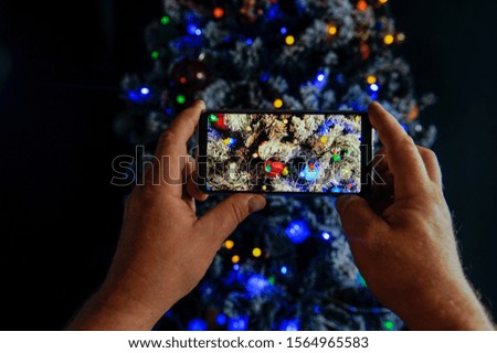 Elderly man holds in his hands new model of  smartphone and take pictures of garlands and Christmas lights on New Year tree. Christmas Eve mixed media 