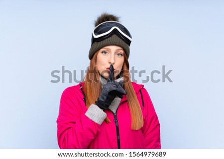 Skier redhead woman with snowboarding glasses over isolated blue wall doing silence gesture