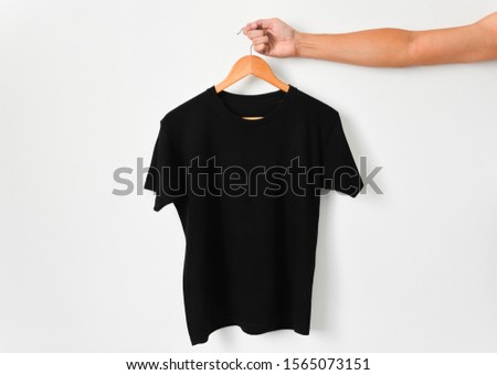 close up of hand holding black color t-shirt hanging on wooden cloth hanger over white color background, copy space
