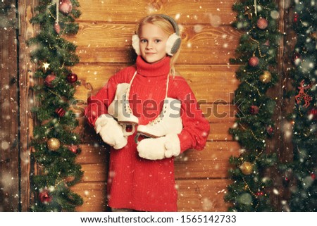 Winter activities. Happy cute girl child in winter clothes is going to skate. She stands under snowfall by a wooden house decorated with Christmas spruce garlands and toys. 