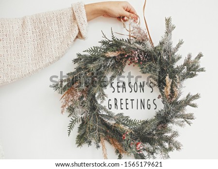 Season's greetings text sign on christmas rustic wreath in girl hand. Creative rural  wreath with fir branches, berries, pine cones, herbs hanging on white wall in room. Seasons greeting card
