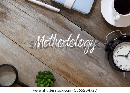 Top view of pen,notebook, a cup of coffee, clock,plant and magnifying glass on wooden background written with Methodology.
