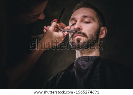 Young gentleman is enjoying mustache and beard trimming at dark barbershop.