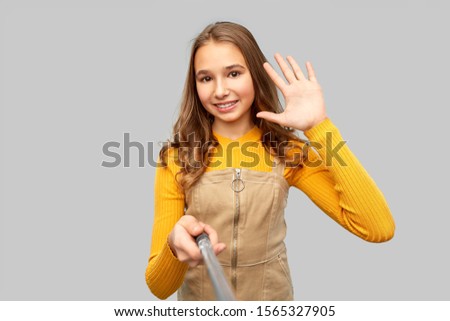 gesture, technology and people concept - happy smiling young teenage girl taking picture by selfie stick and waving hand over grey background