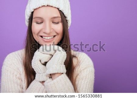 Cute girl with dark hair in white knitted hat and white mittens clasped her hands under her chin. He looks down and smiles broadly. Photo on purple background. The atmosphere of comfort. Winter mood.