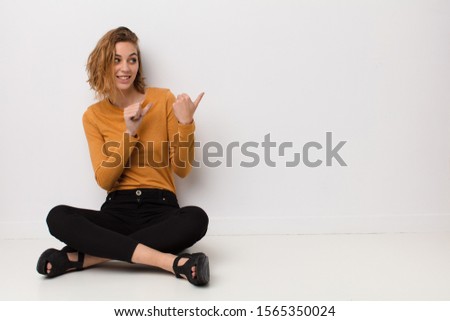 young blonde woman smiling cheerfully and casually pointing to copy space on the side, feeling happy and satisfied against flat color wall