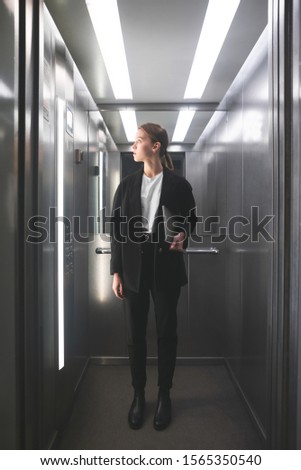 Vertical photo of young calm businesswoman holding laptop in her hand and looking at floor number in lift. Female office worker keeping laptop in her hand looking up in elevator. Full-length photo.