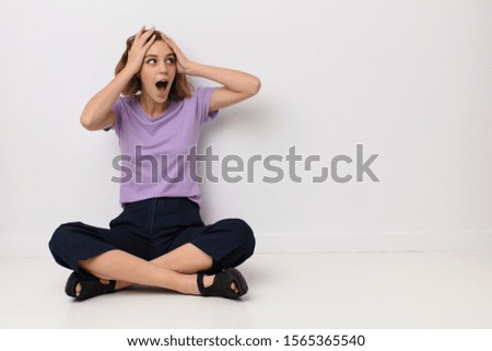young blonde woman with open mouth, looking horrified and shocked because of a terrible mistake, raising hands to head against flat color wall