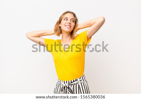 young blonde woman smiling and feeling relaxed, satisfied and carefree, laughing positively and chilling against flat color wall