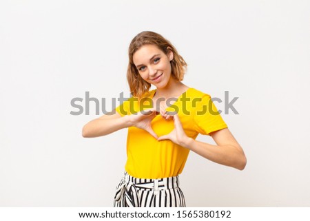 young blonde woman smiling and feeling happy, cute, romantic and in love, making heart shape with both hands against flat color wall