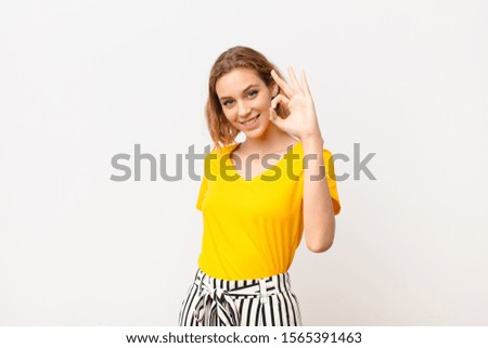 young blonde woman feeling happy, relaxed and satisfied, showing approval with okay gesture, smiling against flat color wall