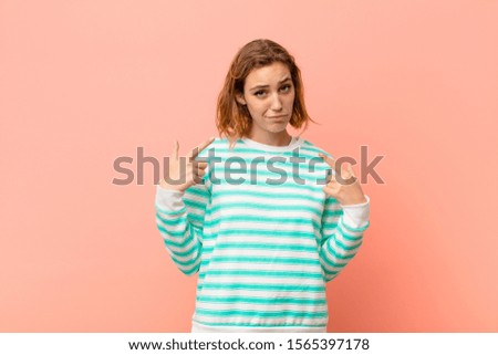 young blonde woman with a bad attitude looking proud and aggressive, pointing upwards or making fun sign with hands against flat color wall