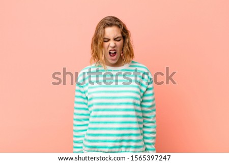 young blonde woman shouting aggressively, looking very angry, frustrated, outraged or annoyed, screaming no against flat color wall