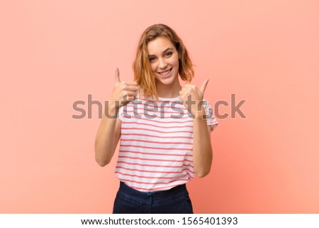young blonde woman smiling joyfully and looking happy, feeling carefree and positive with both thumbs up against flat color wall